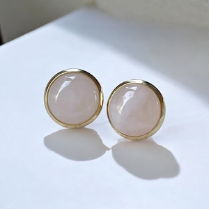 Può includere: Un paio di orecchini a bottone rotondi. Ogni orecchino presenta una pietra rosa pallido lucidata incastonata in una lunetta color oro. Gli orecchini sono esposti su una superficie bianca, con ombre proiettate sotto.