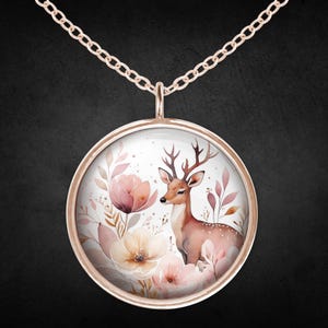 Puede incluir: Un collar de oro rosa con un colgante redondo. El colgante muestra una ilustración de acuarela de un ciervo rodeado de flores y hojas rosas y melocotón. La cadena es delicada y combina con el diseño del colgante.