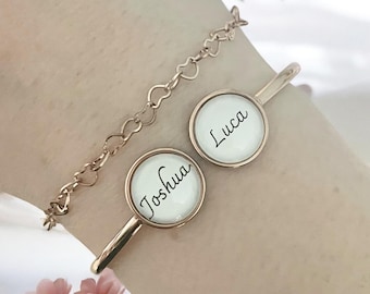 Op de afbeelding: Gepersonaliseerde armbanden in roségoud. Eén armband heeft een delicate hartjesketting. Twee extra armbanden hebben ronde, witte vlakken met de namen "Joshua" en "Luca" in zwart schrift. De armbanden worden getoond op een pols met roze bloemen op de achtergrond.