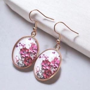 Könnte beinhalten: Ein Paar goldfarbene Ohrhänger. Jeder Ohrring hat einen ovalen Anhänger mit einem Blumendesign in Rosa und Rot auf weißem Hintergrund. Die Ohrringe haben einen Hakenverschluss.