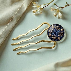 Könnte beinhalten: Goldfarbener Haarkamm mit einem dunkelblauen, kreisförmigen Mittelstück. Das Mittelstück hat ein Blumendesign mit goldenen Akzenten und kleinen Kristallen. Der Kamm hat vier wellenförmige Zinken. Weiße Blumen und cremefarbener Stoff im Hintergrund.