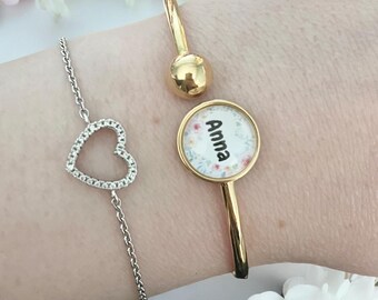 Op de afbeelding: Drie armbanden worden getoond. Een zilveren kettingarmband met een hartvormige hanger bezet met heldere stenen. Een goudkleurige armband met een ronde bedel met de naam "Anna" erop. Een goudkleurige armband met een ronde bal.
