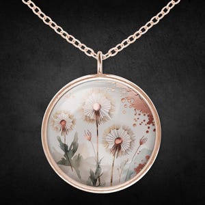 Könnte beinhalten: Eine roségoldene Halskette mit einem runden Anhänger, der ein Aquarellgemälde von Löwenzahnpflanzen zeigt. Der Anhänger hat eine transparente Glasabdeckung, und die Kette ist ebenfalls roségoldfarben. Der Hintergrund verwendet eine weiche, gedämpfte Farbpalette.