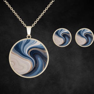 Puede incluir: Un collar dorado con un colgante redondo y pendientes a juego. El colgante y los pendientes presentan un diseño en espiral en tonos azules, blancos y dorados. El conjunto de joyas se muestra sobre un fondo oscuro.