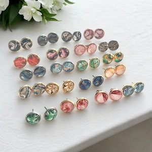 May include: A collection of 18 pairs of round stud earrings with various abstract marbled designs in shades of pink, blue, green, gold, and black. The earrings have gold and silver settings.