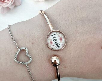 Op de afbeelding: Een roségouden manchetarmband met een cirkelvormige bedel met de naam "Leonie" in zwart schrift. Een zilveren armband met een hartvormige bedel is ook zichtbaar. Roze bloemen zijn op de achtergrond.