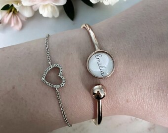 Op de afbeelding: Een zilveren kettingarmband met een hartvormige hanger en een goudkleurige manchetarmband met de naam "Emilia" in script. De manchet heeft een ronde bedel en een bolvormig accent. Zachte roze en witte bloemen op de achtergrond.