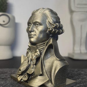 May include: A metallic gold-toned bust sculpture depicting a historical figure with curled hair, wearing a high-collared coat and a cravat. The figure is shown in profile, mounted on a rectangular base with a textured, shimmering finish throughout.