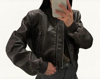 Pode incluir: Casaco bomber castanho escuro com fecho de correr, punhos canelados e um pequeno bolso na manga esquerda. O casaco tem um aspeto ligeiramente envelhecido, semelhante ao couro.