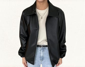 Pode incluir: Casaco de couro preto com gola e fecho de correr. O casaco, com mangas compridas, é usado sobre uma camiseta branca e jeans azuis. Um cinto preto e um colar de prata também são visíveis.