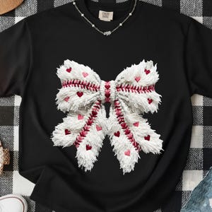 Könnte beinhalten: Schwarzes T-Shirt mit einer großen, strukturierten weißen Schleife mit roten Herzakzenten. Die Schleife hat ein baseballähnliches Stichdesign und kleine rote Herzen. Eine zarte Silberkette ist sichtbar. Das Shirt liegt auf einer schwarz-weiß karierten Oberfläche.