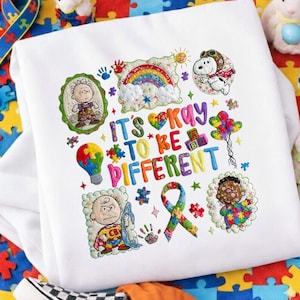 Könnte beinhalten: Weißes Sweatshirt mit farbenfrohem Stickdesign. Mit dem Text "It's Okay to Be Different" mit Bildern eines Regenbogens, Snoopy, Charlie Brown, Puzzleteilen und einem Band. Das Design ist in verschiedenen Farben gehalten.