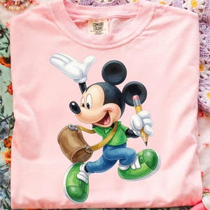Op de afbeelding: Een lichtroze t-shirt met een afbeelding van Mickey Mouse in een groen shirt en blauwe broek, met een bruine tas en een gele potlood in de hand. Het label in de nek bevat de tekst Comfort Colors, 100% cotton.