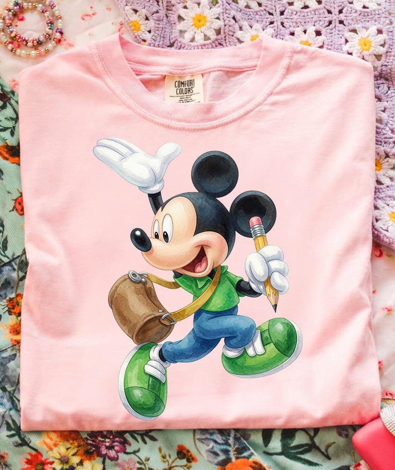 Op de afbeelding: Een lichtroze t-shirt met een afbeelding van Mickey Mouse in een groen shirt en blauwe broek, met een bruine tas en een gele potlood in de hand. Het label in de nek bevat de tekst Comfort Colors, 100% cotton.