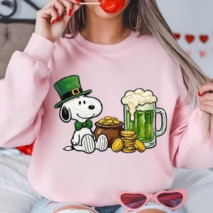 May include: Pink sweatshirt featuring a cartoon Snoopy character wearing a green leprechaun hat and bow tie. Beside Snoopy is a pot of gold coins, a stack of gold coins, and a green beer in a glass mug. The sweatshirt is a light pink color.