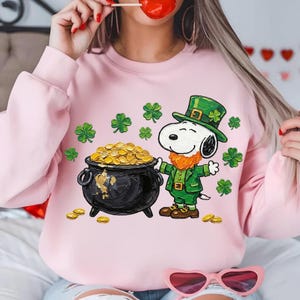 May include: Pink sweatshirt featuring a St. Patrick's Day design. The graphic includes Snoopy dressed as a leprechaun next to a pot of gold and green shamrocks. Gold coins are scattered around the pot. The design is on a pink sweatshirt.