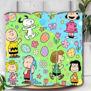 Könnte beinhalten: Drei Edelstahlbecher mit einem Ostermotiv. Das Design zeigt Zeichentrickfiguren, Ostereier, Blumen und Schmetterlinge auf einem grün-blauen Hintergrund. Jeder Becher hat einen silbernen Deckel und einen Strohhalm.