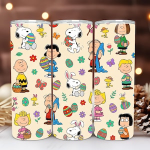 Könnte beinhalten: Drei zylindrische Becher mit einem Ostermotiv mit Snoopy, Charlie Brown und anderen Peanuts-Charakteren. Das Design umfasst Ostereier, Blumen und Schmetterlinge auf cremefarbenem Hintergrund. Die Becher sind aus Edelstahl.