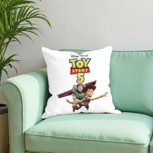 Puede incluir: Un coj&iacute;n cuadrado blanco con el logotipo de Disney Pixar Toy Story 5 en texto azul, amarillo y rojo. Debajo del logotipo, los personajes Woody y Buzz Lightyear aparecen en una pose din&aacute;mica, con Buzz volando y Woody extendiendo la mano.