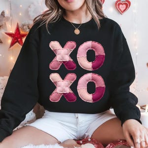 Könnte beinhalten: Schwarzes Sweatshirt mit einem aufgestickten "XOXO"-Design in Rosa- und Burgundertönen. Die Buchstaben haben ein gestepptes Aussehen. Dieses lässige Sweatshirt ist ideal für den Valentinstag oder um Zuneigung auszudrücken.