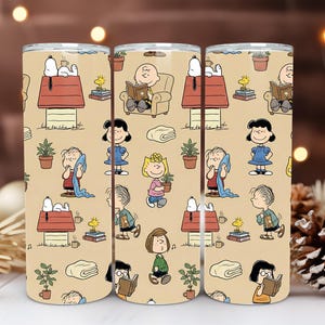 Könnte beinhalten: Drei Edelstahlbecher mit einem sich wiederholenden Muster von Snoopy und der Peanuts-Bande auf beigem Hintergrund. Das Design umfasst Snoopy auf seinem Hundehütte, Charlie Brown beim Lesen und andere Charaktere.