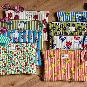 Puede incluir: Una colección de neceseres acolchados y coloridos con varios diseños, incluyendo florales, frutales y a rayas. Cada neceser presenta un lazo a juego y una pequeña etiqueta. Los neceseres son en tonos rojos, verdes, amarillos y azules.