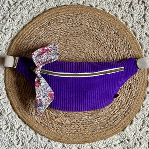 May include: A purple corduroy fanny pack with a white zipper and a floral bow. The bag has a cream-colored adjustable strap and is set on a woven straw mat.