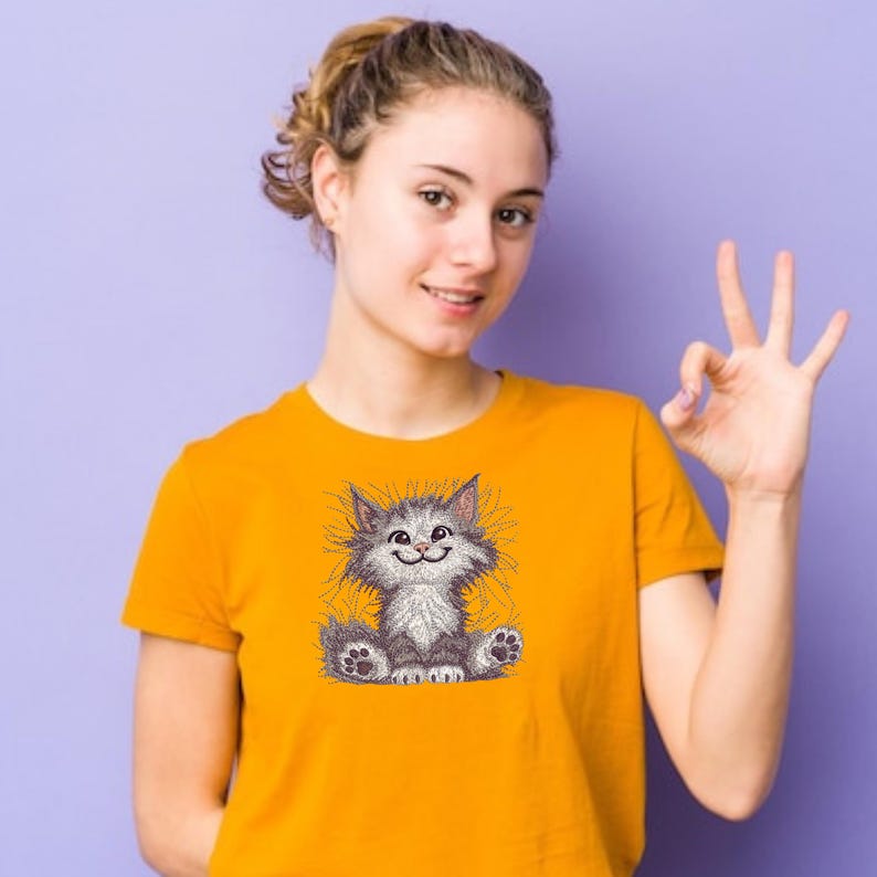 Smiling girl in orange t-shirt showing OK sign, wearing cute fluffy cat embroidery design, perfect for pet lovers and machine embroidery fans.