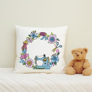 Floral sewing machine embroidery frame stitched on white pillow, placed beside teddy bear in cozy room. Ideal for nursery, gifts, or handmade decor