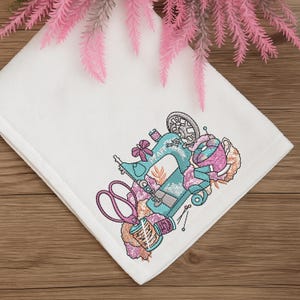 White towel decorated with sewing machine embroidery design featuring scissors, thread, and sewing motifs – digital download embroidery file for towels, linens, DIY sewing, and machine embroidery home projects