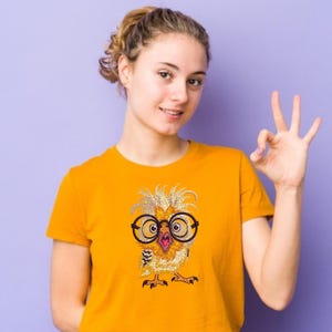 Smiling girl wearing bright yellow t-shirt with cute owl embroidery design featuring large glasses, shown on purple background – perfect for fun American embroidery projects.