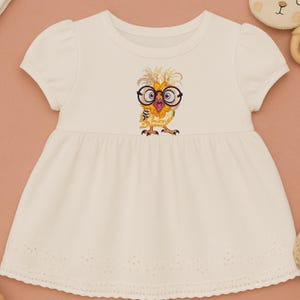 White baby dress with cute owl embroidery design featuring glasses, flat lay styled with neutral soft toys on peach background – perfect for baby shower gifts and newborn outfits in USA and Europe.