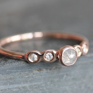 May include: A delicate rose gold band ring with five bezel-set diamonds. The center diamond is larger than the others and has a slightly cloudy appearance.