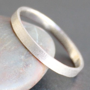 May include: A close-up of a silver ring with a brushed finish. The ring is round and appears to be made of metal. The ring is resting on a light-colored, textured surface.