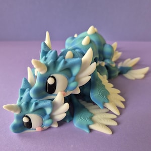 Puede incluir: Figura de dragón impresa en 3D en tonos azules y blancos. El dragón tiene ojos grandes y expresivos, cuernos blancos y estructuras similares a alas. La figura está tumbada sobre una superficie morada.