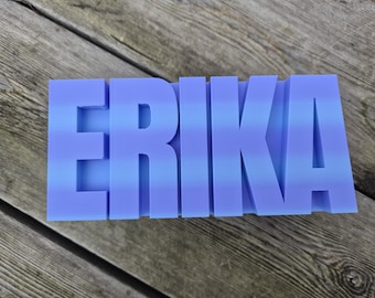 May include: A blue and purple ombre-colored name sign that spells out the name "ERIKA". The letters are thick and blocky, and the sign is resting on a weathered wooden surface.