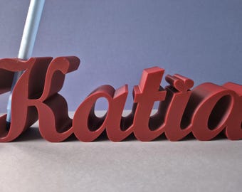 May include: A maroon-colored, 3D-printed name, "Katia", in a cursive font. The letters are thick and stand upright. A white pen is inserted into the letter "K". The background is a gradient of light gray and blue.