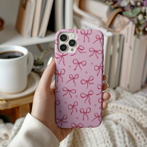 Puede incluir: Una funda de teléfono rosa con un patrón repetido de lazos rojos y pequeños corazones. La funda tiene un acabado liso y está diseñada para un teléfono inteligente. La funda está en la mano de una persona.