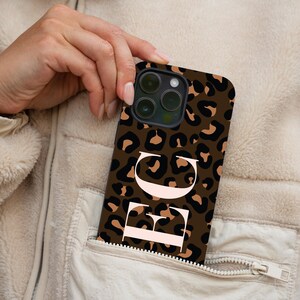 Peut inclure: Coque de téléphone marron avec un motif léopard et les lettres blanches "EG". L'étui est tenu dans une poche d'une veste beige.