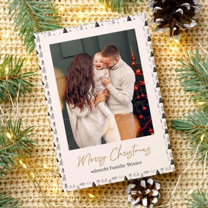 May include: A Christmas card with a family photo inside, surrounded by a decorative border of small black and white trees. The words "Merry Christmas" are written in gold script, with additional text below. The card is set against a backdrop of pine needles and a knitted blanket.