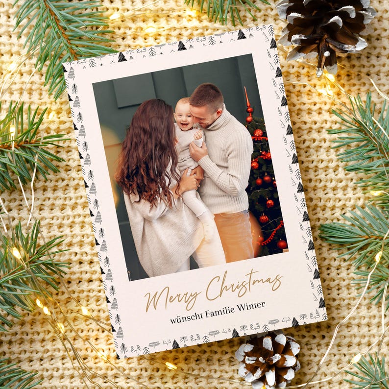May include: A Christmas card with a family photo inside, surrounded by a decorative border of small black and white trees. The words "Merry Christmas" are written in gold script, with additional text below. The card is set against a backdrop of pine needles and a knitted blanket.