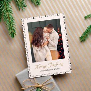 May include: A Christmas card featuring a family portrait with the words "Merry Christmas" in gold script. The card has a decorative border with small black trees. A wrapped gift and pine branches are also visible.
