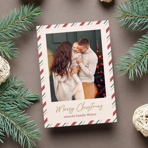 May include: A Christmas card featuring a family portrait with a festive border. The card has a cream background with a red and green striped border. The text "Merry Christmas" is written in gold script, with additional text below. The card is surrounded by pine branches and decorative balls.