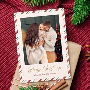 May include: Christmas card featuring a family with a baby, framed with a red and green striped border. The card reads "Merry Christmas" in gold script, with additional text below. The card is placed on a wrapped gift.