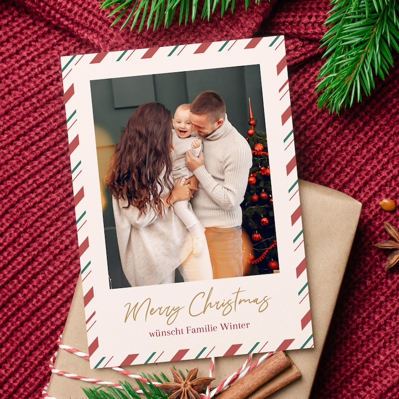 May include: Christmas card featuring a family with a baby, framed with a red and green striped border. The card reads "Merry Christmas" in gold script, with additional text below. The card is placed on a wrapped gift.