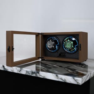 May include: A wooden watch winder with a clear glass door and two watch holders. One watch has a black band and silver face, the other has a green face. The winder sits on a marble countertop.