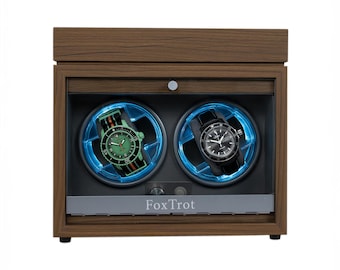 May include: A wooden watch winder with two compartments, each illuminated with blue light and holding a wristwatch. One watch has a green face and striped band, the other a black face and band. The name "FoxTrot" is printed on the front.