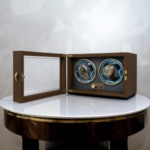 May include: A wooden watch winder with two compartments, each holding a wristwatch. The winder has a glass door and a control panel. The watches are displayed against a dark background, illuminated by blue lights.