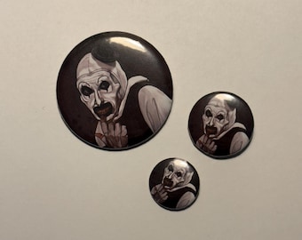 May include: Three circular buttons with a horror theme. Each button displays a black background with a white and grey illustration of a character with white face paint, a white head covering, and a black vest. The buttons are different sizes.