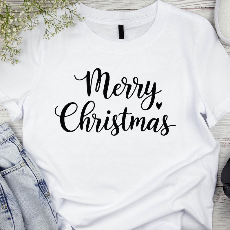 May include: White t-shirt with the words "Merry Christmas" in black script. A small black heart is next to the word "Christmas". The shirt is made of a soft material and is perfect for the holiday season.